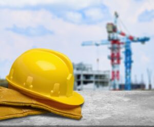 Yellow safety helmet and work gloves resting on a concrete surface, with a construction site featuring cranes and unfinished buildings blurred in the background.