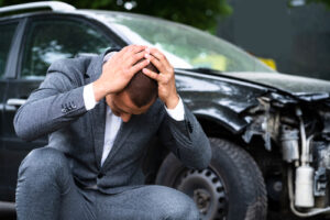 Distressed man in a business suit sitting beside a damaged car, holding his head in despair, highlighting the emotional impact of a vehicular accident.