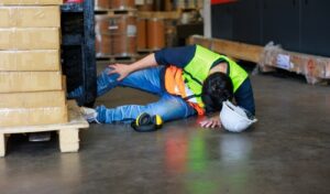 A worker wearing a safety vest and helmet has fallen next to a pallet in a warehouse, illustrating a workplace accident and the importance of safety measures.