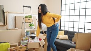 A woman in a yellow sweater and blue jeans stands in a cluttered living room surrounded by boxes, gripping her lower back and wincing in pain, indicative of a back injury possibly due to moving or lifting heavy objects.