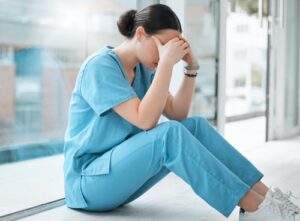 A female healthcare worker in blue scrubs sits on the floor, leaning against a large glass window. She appears distressed or overwhelmed, covering her face with her hands. The setting suggests a break or a moment of exhaustion in a medical facility.