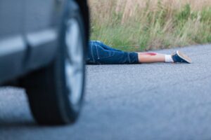 A boy lies on the roadway behind a parked car, visibly injured with wounds on his legs.