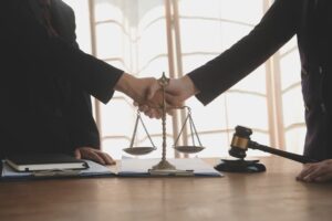 A lawyer shaking hands with a female client at their desk, surrounded by documents, contracts, and a gavel on the table, symbolizing legal agreements and cooperation.