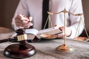 A male lawyer or judge sitting at a desk in an office, working with law books, a gavel, and scales of justice while reviewing a case report.