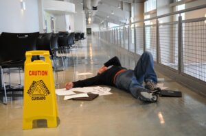 A man lying on a wet floor after slipping, with a caution sign visible in the background. The focus is on the man's head.