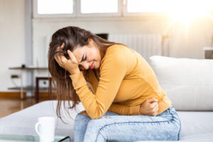 A woman sitting on a couch, holding her head in pain with a pained expression, appearing distressed while clutching her stomach, with a cup nearby.