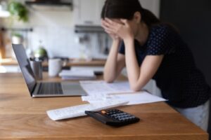 A woman sitting at a wooden table, holding her head in her hands, appearing stressed while looking at bills and a laptop. A calculator is in the foreground.