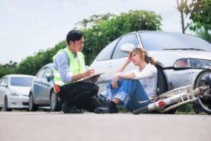 A concerned man in a safety vest takes notes while assisting an injured woman sitting on the ground next to a damaged bicycle and car.