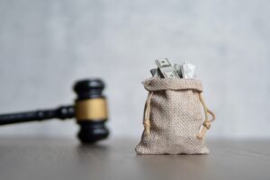 A burlap sack filled with cash sits on a wooden surface next to a judge's gavel, symbolizing financial compensation or legal settlements.