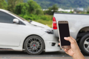 A person holds a smartphone, taking a photo of a rear-end collision between a white car and a pickup truck for documentation.