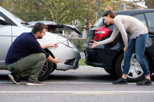 A man and a woman examine the damage on their vehicles after a rear-end collision, discussing the accident on the roadside.