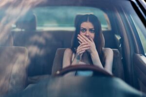 A young woman with a fearful expression covering her mouth inside a car, conveying anxiety, emotional distress, and concern against a blurred background.