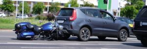 A close-up view of a motorcycle crashed into the back of a car on the road. The image highlights the point of impact, showing visible damage to both vehicles.