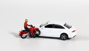 A miniature motorcycle rider is positioned in front of a white miniature car, depicting the concept of a dangerous motorcycle accident. The scene emphasizes the risks motorcyclists face on the road.